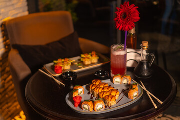 Set of various delicious mouth-watering sushi rolls on the table with a cocktail, soy sauce, chopsticks and a vase with a flower in the restaurant in the evening, a selective focus.