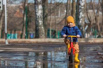 A cheerful little boy rides a bike through puddles. A happy child walks outside in the spring. The kid is dressed in a fashionable plaid shirt, overalls and yellow rubber boots.