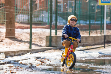 A cheerful little boy rides a bike through puddles. A happy child walks outside in the spring. The kid is wearing a fashionable plaid shirt, sunglasses and yellow rubber boots.