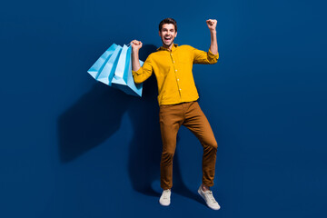 Full size photo of handsome young man raise fists shopping bags promo wear trendy yellow outfit isolated on dark blue color background