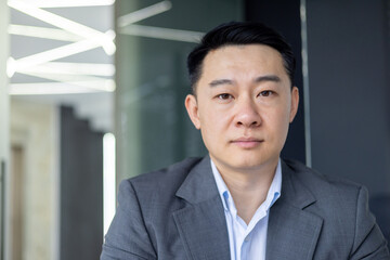 A professional Asian man in business attire stands confidently in an office, exuding leadership and determination.