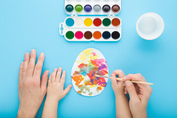 Mother and baby boy hands holding paintbrush and painting colorful egg on paper with watercolor on blue table background. Closeup. Child making easter decoration. Point of view shot. Top down view. - 773063311