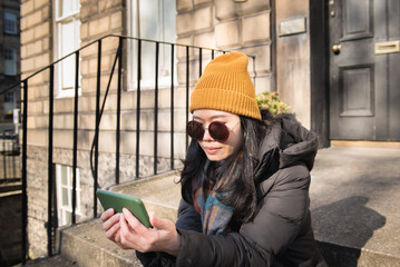 Young Asian woman wearing  yellow hat watches content on her green mobile phone in a sunny day while sitting on the steps at the front door of a house in Edinburgh, Scotland, UK