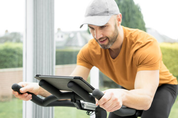 Fototapeta na wymiar Young Hispanic man with a stubble wearing a grey cap and orange top exercising on a spinning bike in a conservatory while following an online fitness workout on his tablet resting on the handlebar
