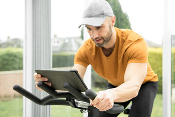 Spanish man with a stubble wearing a grey cap and orange top exercising on a spinning bike in a...