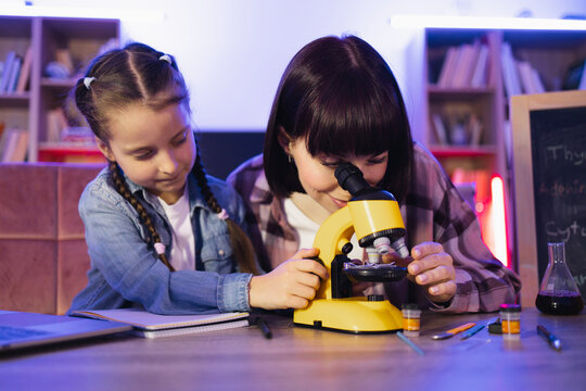 Focused adult mother looking at onion cells in electron microscope while her daughter sitting near her and studying process. Concept of family and time together at evening time.