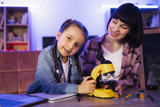 Focused little girl looking camera, working with electron microscope while her mother sitting near and controlling process. Concept of family and time together at evening time.