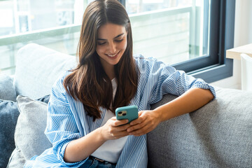 Happy girl checking social media while holding smartphone at home. Smiling young brunette woman...