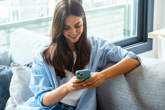 Happy girl checking social media while holding smartphone at home. Smiling young brunette woman using mobile phone app to play games, shop online, order delivery while relaxing on sofa. Scrolling.