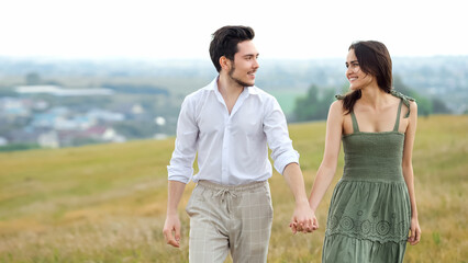 Happy couple of young man and woman smiles walking by hand across field on hilly plain. Lovers joy and landscape against city