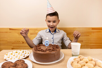 A boy is sitting in front of a table with a birthday cake, in which he blows out a candle in the form of the number 8. The concept of a birthday celebration.