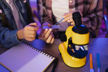 Close up of hands of young woman in casual wear sitting with girl at table and using microscope for studying sciences. Mother with daughter holding slides with plant cell near microscope at home.