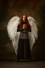 Red-haired maiden in angelic wings and medieval attire embodies a serene, prayerful stance against...