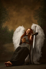 Redhead woman looks as angel in chainmail with sword, with large white wings and golden halo...