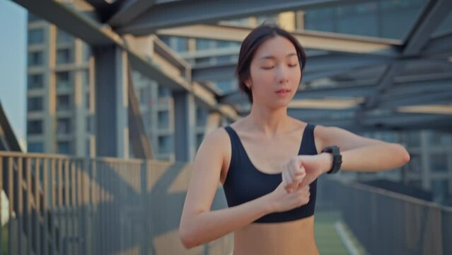 Young asian woman runner is training and checking her smart watch in the summer sunset within a city, The athlete fit and firm girl exercising by jogging workout sport outdoor for her healthy wellness