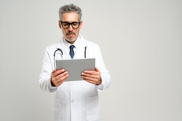 Senior doctor with grey hair is deeply focused on tablet, reviewing patient information or medical...