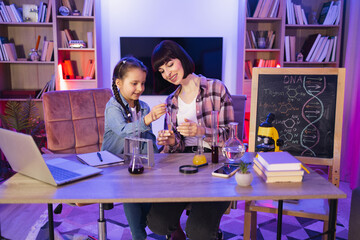 Parent mother with daughter kid making chemical test at evening time at home living room. Scientific experiment at home with nanny, babysitter or teacher. Laboratory tests for school homework.