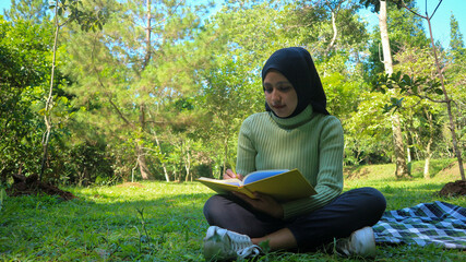 Relaxed muslim woman enjoying weekend at park, sitting on grass and writing book, empty space