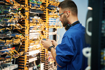 Holding smartphone. Young man is working with internet equipment and wires in server room