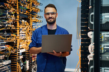 With laptop in hands. Young man is working with internet equipment and wires in server room