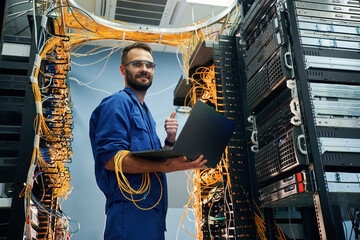 Silver colored laptop in hands. Young man is working with internet equipment and wires in server...