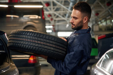 Holding a tire. Car repairman is in the garage with automobile