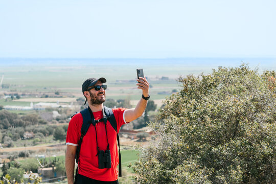 A man is taking a picture of himself and the landscape with his cell phone while wearing a red shirt and a black hat on a natural trail