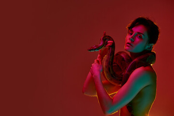 Portrait of sensual, beautiful young woman in slim body in underwear standing with snake against red background in neon light. Passion. Concept of female beauty, animal theme, care, elegance, fashion