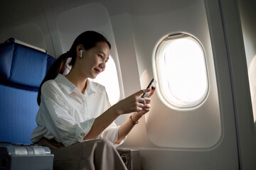 Young Asian woman uses mobile phone to check news information Sitting near the window in business class, airplane class during flight, travel and business concept
