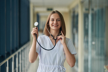Using stethoscope. Female doctor in white coat is in the hall