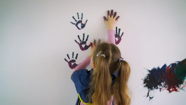 Back view of a fun mischievous baby girl leaving colored hand prints on white wall wall in room. Children's palms dipped in paint make prints on the wall. High quality 4k footage
