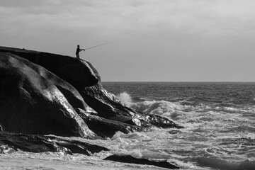 Black and white of a person standing on a rocky shoreline, fishing in the calm waters.