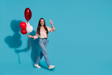 Fototapeta premium Full length photo of lovely girl wear silk blouse hold heart balloons go empty space show v-sign isolated on blue color background