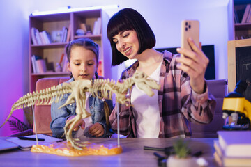 Young woman and little cute girl filming video of their scientific research. Happy mother examine skeleton of dinosaur with smart cute daughter recording content for natural blog.