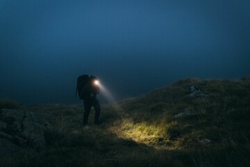 Young adult male walking atop a hill against a night sky, wearing a backpack © Wirestock