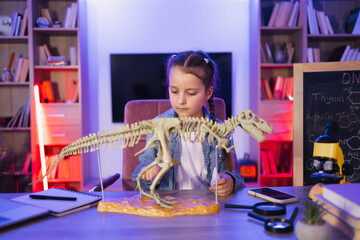 Portrait of smiling little girl sitting at table and examine skeleton of dinosaur. Caucasian child in casual wear making model of tyrannosaurus out of bones in evening at home.