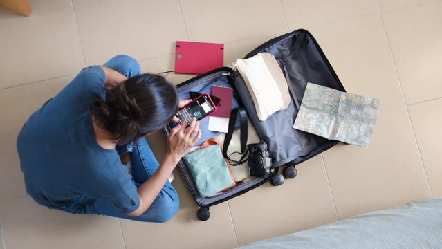 Woman preparing suitcase for trip alone. Checking suitcase and mobile application, flights, trip route, destination, and documentation. Camera passports and clothes. Young woman traveling alone.
