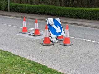 The edge of a manhole cover has broken away forming a pothole in the road and cones and an arrow sign are in place to divert the traffic