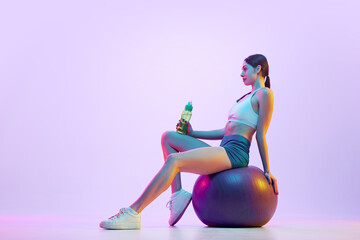 Young woman in sportswear posing sitting on fit-ball with battle of water in neon light against...