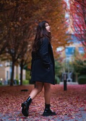 Vertical shot of a young female in a coat posing in a park covered with autumn foliage and trees