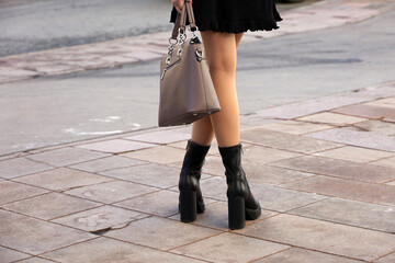 Female legs in black boots on high heels. Slim girl in short skirt and pantyhose standing with purse on a street, fashion in spring city