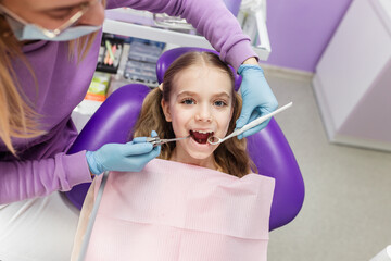 Portrait of a cute little girl laughing looking at camera sitting in stomatology seat while pediatric stomatologist is ready to do teeth examination