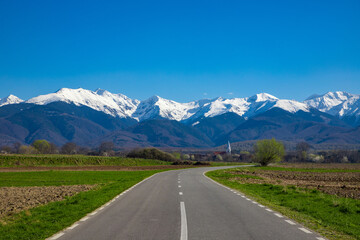 Fototapeta na wymiar Landscape with a road leading to a village and in the background the Fagaras mountains with their peaks covered with snow. Perfect for wallpaper or design. Beautiful landscape from Romania