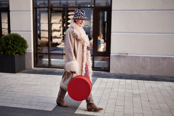 Fashionable mature woman walking in the city, dressed in a long beige coat, Cossack boots, hat with animal print and original handbag box. Street style trend for mature women