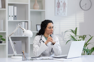 Thoughtful Hispanic female doctor sitting at her desk in a bright medical office, looking at a laptop screen.