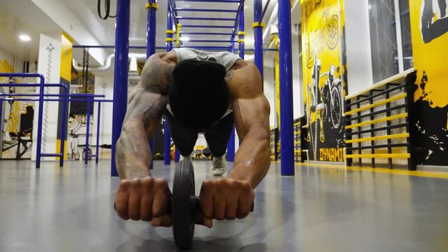 Strong black guy trying to do exercises with gymnastic wheel at modern gym. Muscular african sportsman practicing work with ab roller at fitness centre. Young afro american athlete training indoor