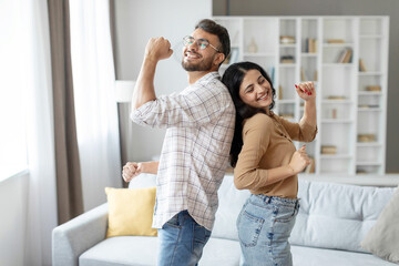 Joyful indian couple dancing in living room interior, having fun and singing, husband and wife...