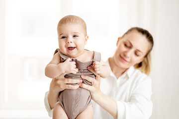 a young mother holds her little newborn baby at arm's length in the bedroom by the window, the...