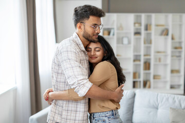 Content indian lady embraces husband, both smiling, sharing warm moment at home, standing in living room interior