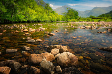 Glen Nevis, Scotland: A tranquil river flows through verdant landscapes, framed by towering mountains under a clear sky.